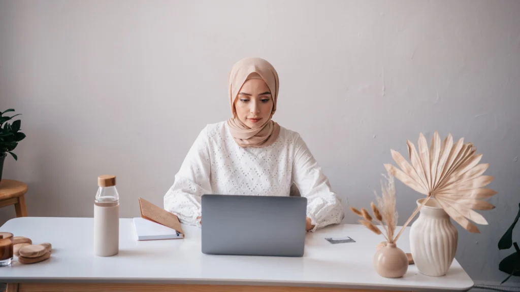 A hijab-wearing woman working on a laptop with a book, bottle, and flower vase nearby, representing a freelance digital marketer in Calicut.