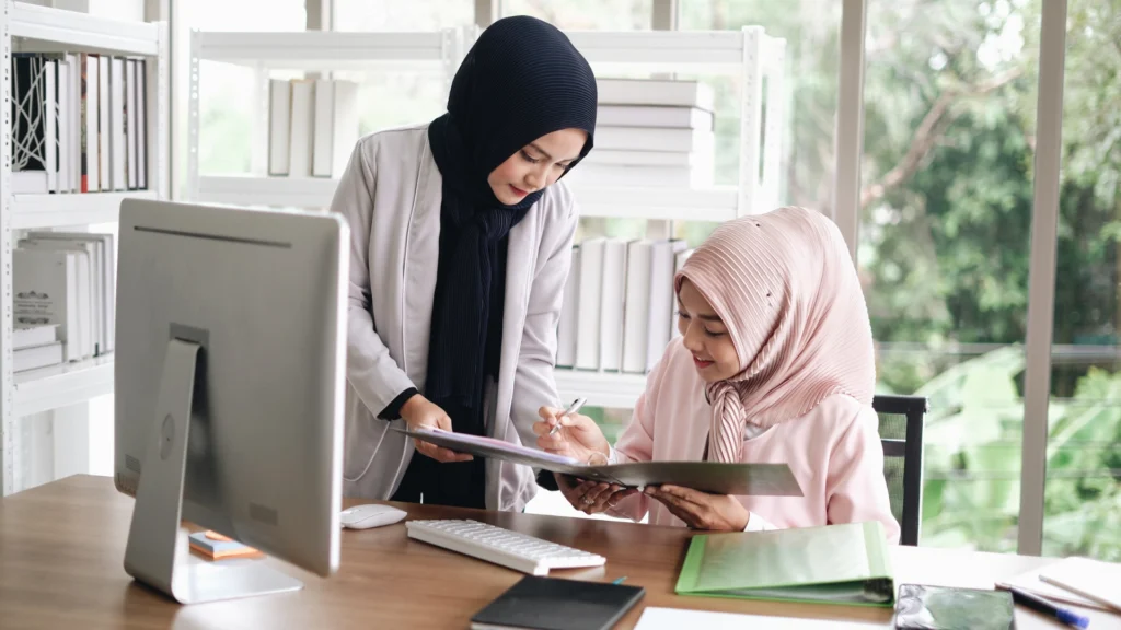 Two hijab-clad women engaged in conversation near a table with a laptop and study tools, symbolizing collaboration as freelance digital marketers in Calicut.